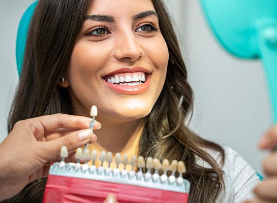 Woman with brown hair smiling into mirror with shade guide held to her teeth