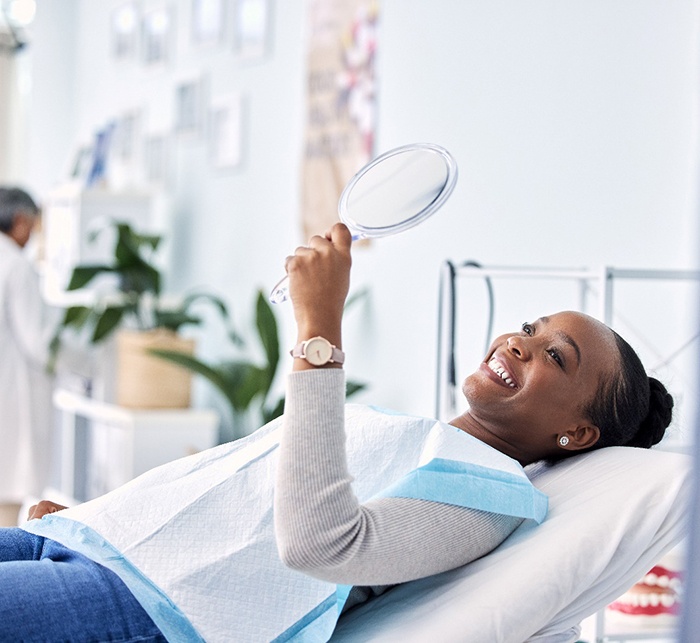 Woman in gray sweater in dental chair smiling at reflection in handheld mirror