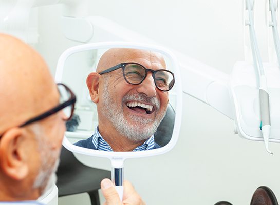 Man with glasses checking smile in mirror at dentist’s office