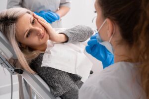 a patient sits in dental exam chair holding the side of her mouth in pain as the dentist lets her know that she will need emergency teeth removal