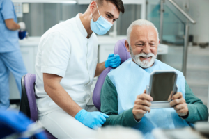 an elder man sits in a dental exam chair holding up a mirror as a dental professional explains and shows the patient examples of restorative dentistry