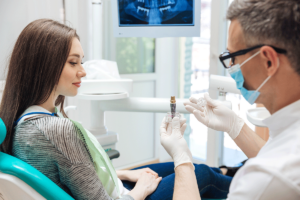 a woman patient sits in an exam chair while her dentist shows her the dental implant procedure while also explaining the pros and cons of dental implants