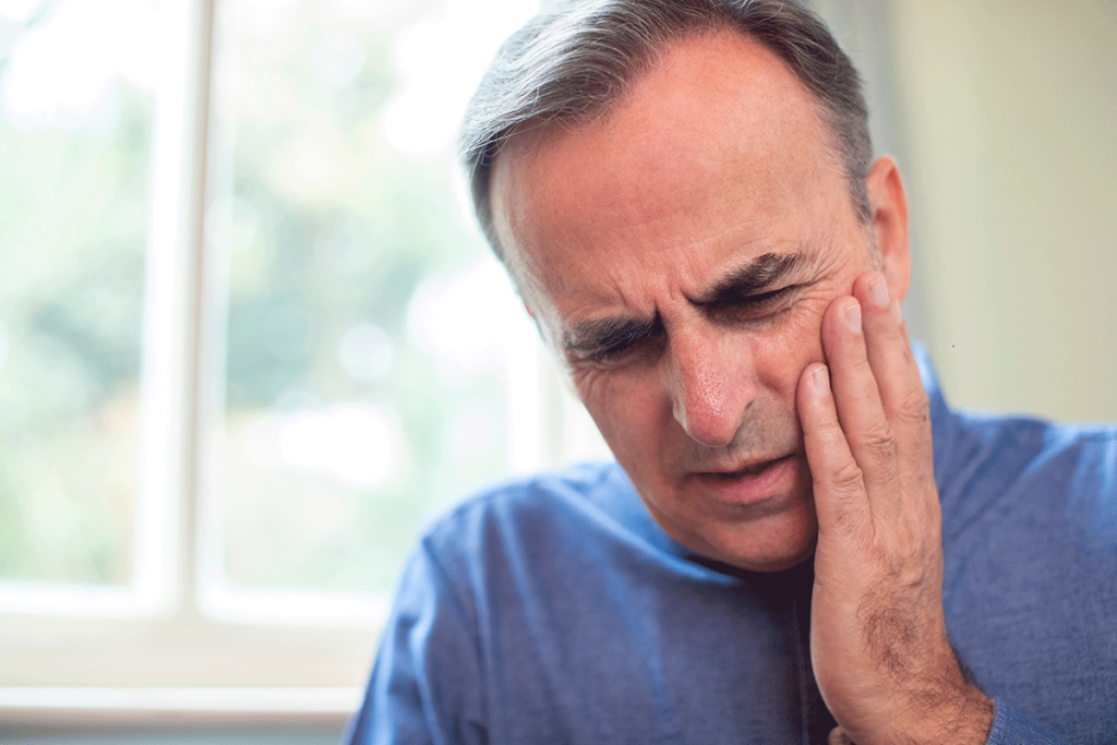 an older adult male holds his hand to the side of his face in pain and wondering what to do in a dental emergency