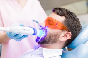 a man sits in a dental exam chair while a dental professional works on whitening yellow teeth of the patient