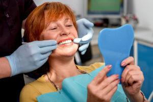 a woman smiles while in a dental exam chair and looking at a mirror while dental professional explains the positive effects of dental implants