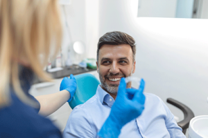 man smiles in exam chair at his appointment for invisalign services while a dental professional stands and holds his invisalign aligners 